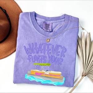 Puede incluir: Una camiseta violeta Comfort Colors con la frase "Whatever Floats Your" y un gr&aacute;fico de un pont&oacute;n. Tambi&eacute;n hay un collar dorado y un sombrero marr&oacute;n en la imagen. La camiseta es de cuello redondo y tiene una etiqueta en el cuello.