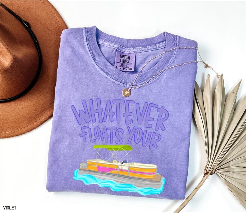 Puede incluir: Una camiseta violeta Comfort Colors con la frase "Whatever Floats Your" y un gr&aacute;fico de un pont&oacute;n. Tambi&eacute;n hay un collar dorado y un sombrero marr&oacute;n en la imagen. La camiseta es de cuello redondo y tiene una etiqueta en el cuello.