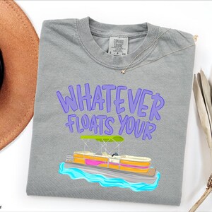 Puede incluir: Camiseta gris con la frase "WHATEVER FLOATS YOUR" en letras moradas. Debajo, una colorida ilustraci&oacute;n de un barco pont&oacute;n sobre agua azul. La camiseta es de la marca Comfort Colors.