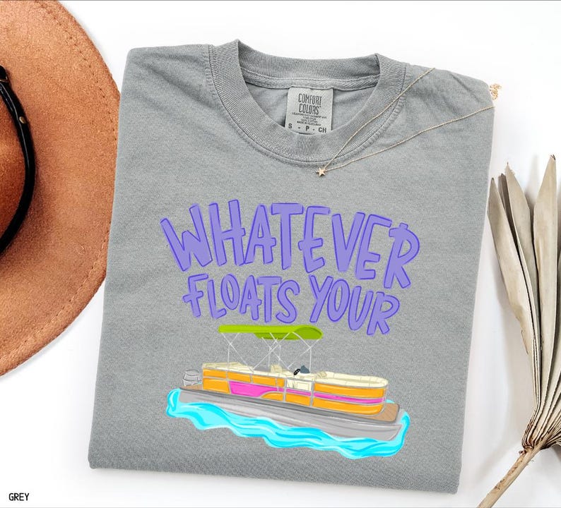 Puede incluir: Camiseta gris con la frase "WHATEVER FLOATS YOUR" en letras moradas. Debajo, una colorida ilustraci&oacute;n de un barco pont&oacute;n sobre agua azul. La camiseta es de la marca Comfort Colors.