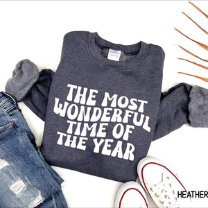 May include: A heather navy crewneck sweatshirt featuring the white text THE MOST WONDERFUL TIME OF THE YEAR in a bold, rounded font. The long-sleeved garment is displayed alongside blue distressed denim jeans and white canvas sneakers.
