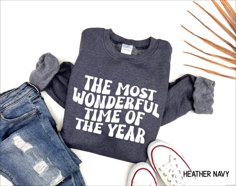 May include: A heather navy crewneck sweatshirt featuring the white text THE MOST WONDERFUL TIME OF THE YEAR in a bold, rounded font. The long-sleeved garment is displayed alongside blue distressed denim jeans and white canvas sneakers.