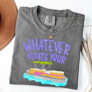 Puede incluir: Una camiseta gris Comfort Colors con la frase "WHATEVER FLOATS YOUR" en letras moradas. Un gr&aacute;fico colorido de un pont&oacute;n est&aacute; debajo del texto. Un collar plateado con un colgante est&aacute; sobre la camiseta.