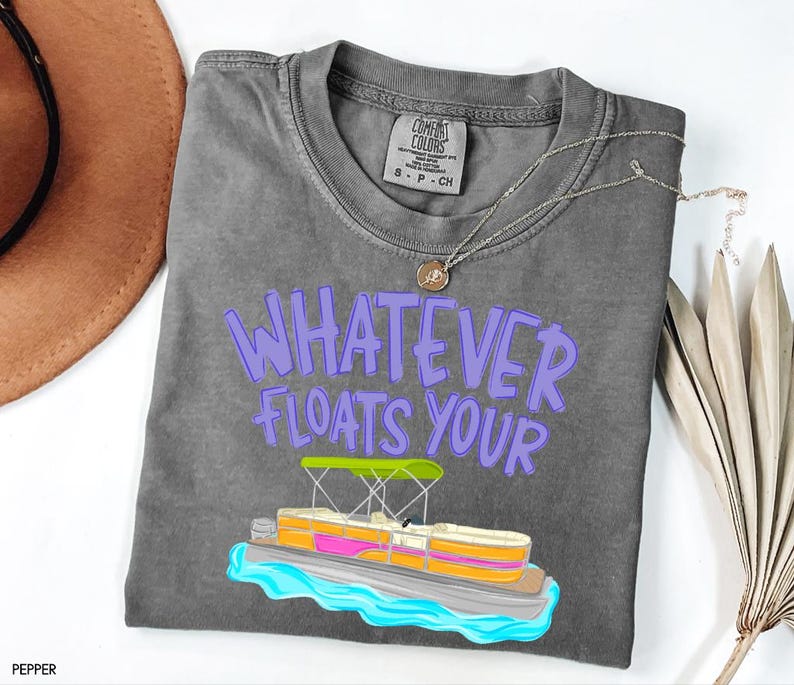 Puede incluir: Una camiseta gris Comfort Colors con la frase "WHATEVER FLOATS YOUR" en letras moradas. Un gr&aacute;fico colorido de un pont&oacute;n est&aacute; debajo del texto. Un collar plateado con un colgante est&aacute; sobre la camiseta.