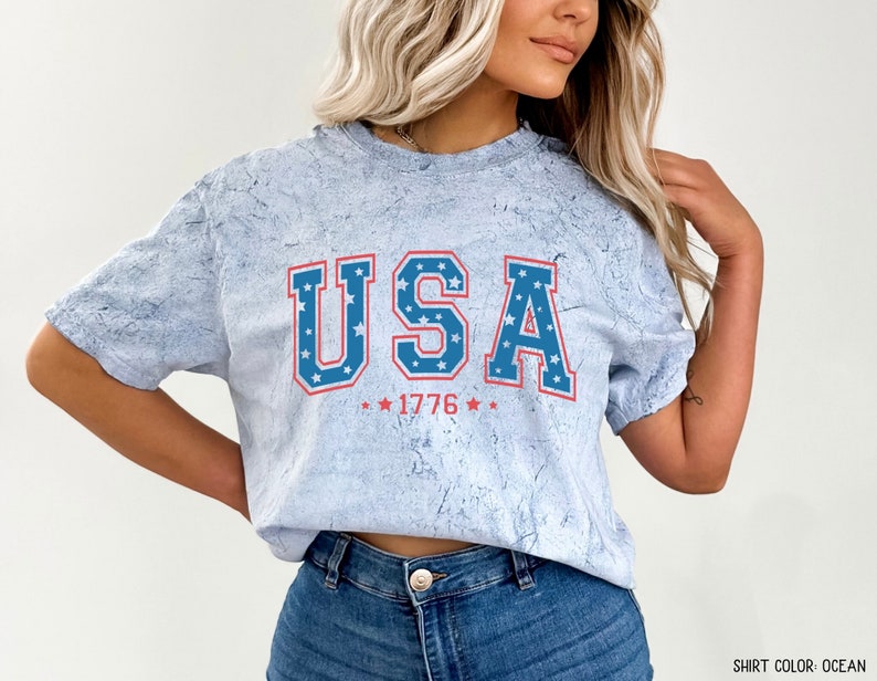 May include: A light blue t-shirt with a distressed look featuring a red, white and blue "USA" graphic with stars and the year "1776".