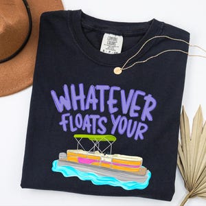 Puede incluir: Camiseta negra con la frase "WHATEVER FLOATS YOUR" en letras moradas y un gr&aacute;fico de un barco pont&oacute;n de dibujos animados. Un collar dorado y un sombrero marr&oacute;n tambi&eacute;n est&aacute;n en la imagen. La camiseta es de un material suave.