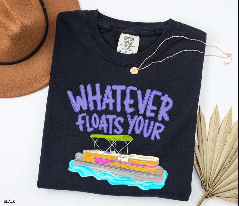 Puede incluir: Camiseta negra con la frase "WHATEVER FLOATS YOUR" en letras moradas y un gr&aacute;fico de un barco pont&oacute;n de dibujos animados. Un collar dorado y un sombrero marr&oacute;n tambi&eacute;n est&aacute;n en la imagen. La camiseta es de un material suave.