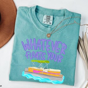 Puede incluir: Camiseta verde mar con la frase "WHATEVER FLOATS YOUR" en letras moradas. Un dibujo animado de un pont&oacute;n est&aacute; debajo del texto. Un collar dorado est&aacute; sobre la camiseta.