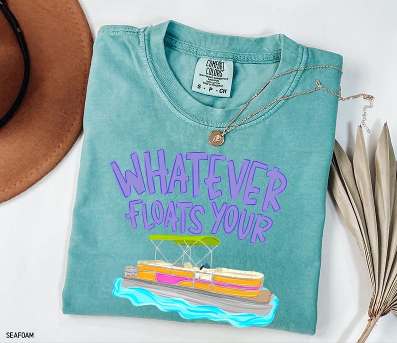 Puede incluir: Camiseta verde mar con la frase "WHATEVER FLOATS YOUR" en letras moradas. Un dibujo animado de un pont&oacute;n est&aacute; debajo del texto. Un collar dorado est&aacute; sobre la camiseta.