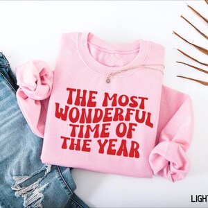 May include: A light pink crewneck sweatshirt featuring the red text THE MOST WONDERFUL TIME OF THE YEAR in a wavy font. The sweatshirt is positioned next to a pair of blue denim jeans with distressed details and a dried palm leaf frond.