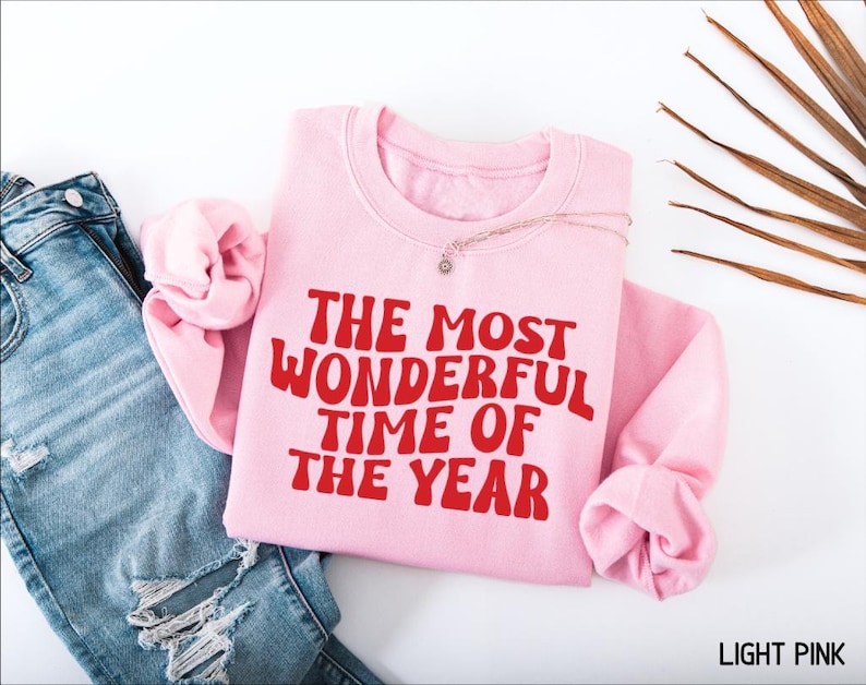 May include: A light pink crewneck sweatshirt featuring the red text THE MOST WONDERFUL TIME OF THE YEAR in a wavy font. The sweatshirt is positioned next to a pair of blue denim jeans with distressed details and a dried palm leaf frond.