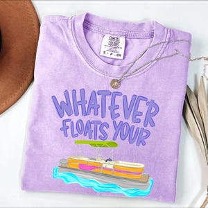 Puede incluir: Una camiseta de color lila claro con la frase "WHATEVER FLOATS YOUR" en morado. Debajo, un dibujo de una lancha pont&oacute;n con detalles rosas, naranjas y amarillos. Un collar plateado con un colgante descansa sobre la camiseta.