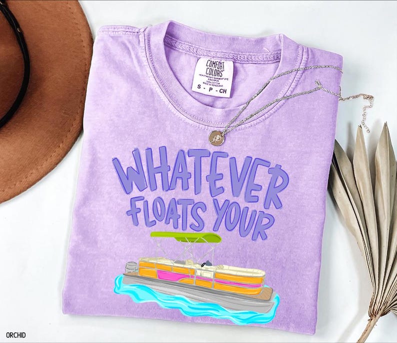 Puede incluir: Una camiseta de color lila claro con la frase "WHATEVER FLOATS YOUR" en morado. Debajo, un dibujo de una lancha pont&oacute;n con detalles rosas, naranjas y amarillos. Un collar plateado con un colgante descansa sobre la camiseta.