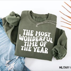 May include: An olive green crewneck sweatshirt featuring the white text "THE MOST WONDERFUL TIME OF THE YEAR" in a wavy, retro-inspired font. The garment is displayed alongside a pair of light blue distressed denim jeans and a silver chain necklace.