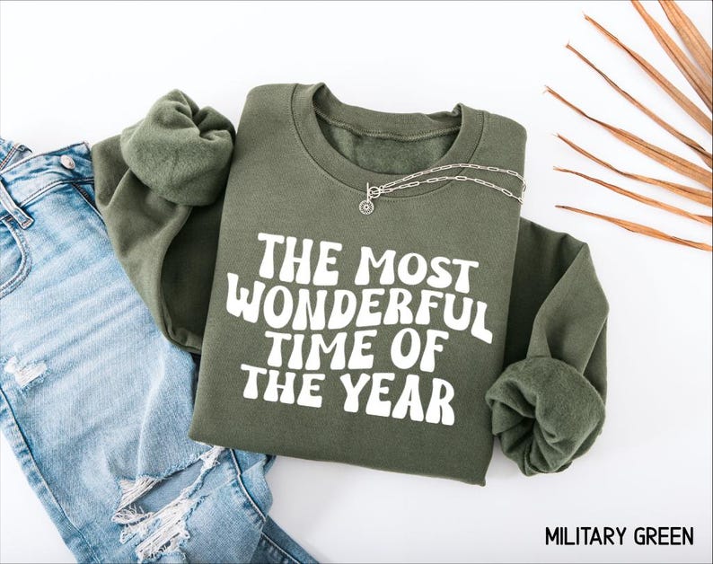 May include: An olive green crewneck sweatshirt featuring the white text "THE MOST WONDERFUL TIME OF THE YEAR" in a wavy, retro-inspired font. The garment is displayed alongside a pair of light blue distressed denim jeans and a silver chain necklace.