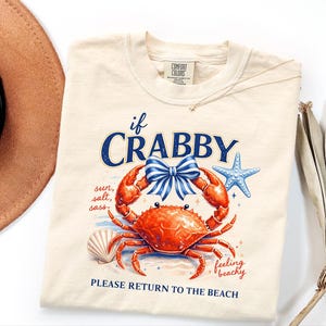 May include: Ivory-colored t-shirt featuring a vibrant orange crab with a blue bow, starfish, and seashell graphic. The text reads "if CRABBY, sun, salt, sass, feeling beachy, PLEASE RETURN TO THE BEACH."