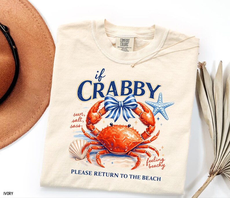 May include: Ivory-colored t-shirt featuring a vibrant orange crab with a blue bow, starfish, and seashell graphic. The text reads "if CRABBY, sun, salt, sass, feeling beachy, PLEASE RETURN TO THE BEACH."