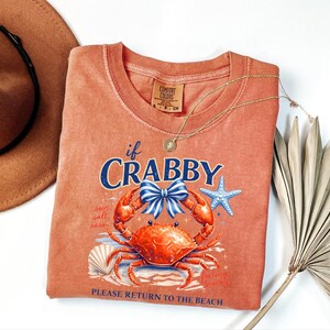 May include: Coral-colored Comfort Colors t-shirt with a crab graphic and the text "if Crabby." The crab has a blue bow, and the design includes a starfish and seashell. A gold necklace and a brown hat are also in the image.