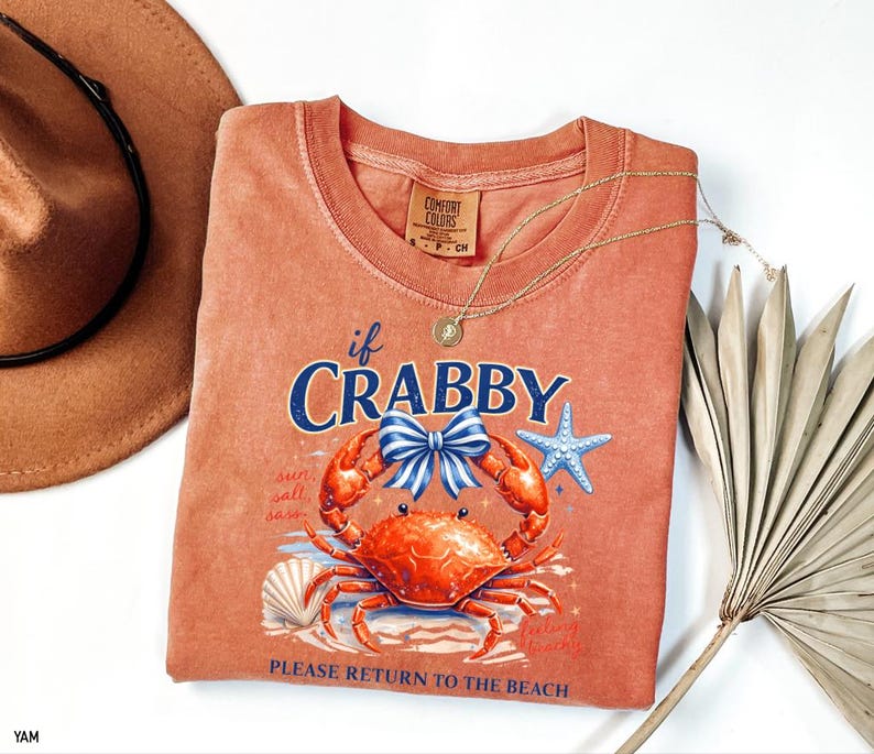 May include: Coral-colored Comfort Colors t-shirt with a crab graphic and the text "if Crabby." The crab has a blue bow, and the design includes a starfish and seashell. A gold necklace and a brown hat are also in the image.