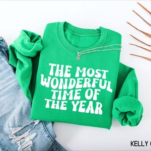 May include: A kelly green crewneck sweatshirt featuring the white text THE MOST WONDERFUL TIME OF THE YEAR in a bold, wavy font. The garment is displayed alongside a pair of distressed blue denim jeans and a silver chain necklace.