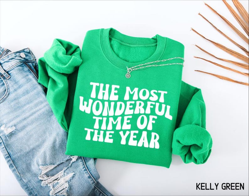 May include: A kelly green crewneck sweatshirt featuring the white text THE MOST WONDERFUL TIME OF THE YEAR in a bold, wavy font. The garment is displayed alongside a pair of distressed blue denim jeans and a silver chain necklace.