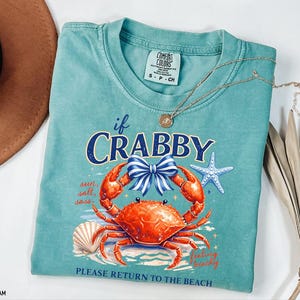 May include: Seafoam green t-shirt with a crab graphic and the text "If Crabby, Please Return to the Beach." The graphic includes a crab, bow, starfish, and seashell. A gold necklace is draped across the shirt.