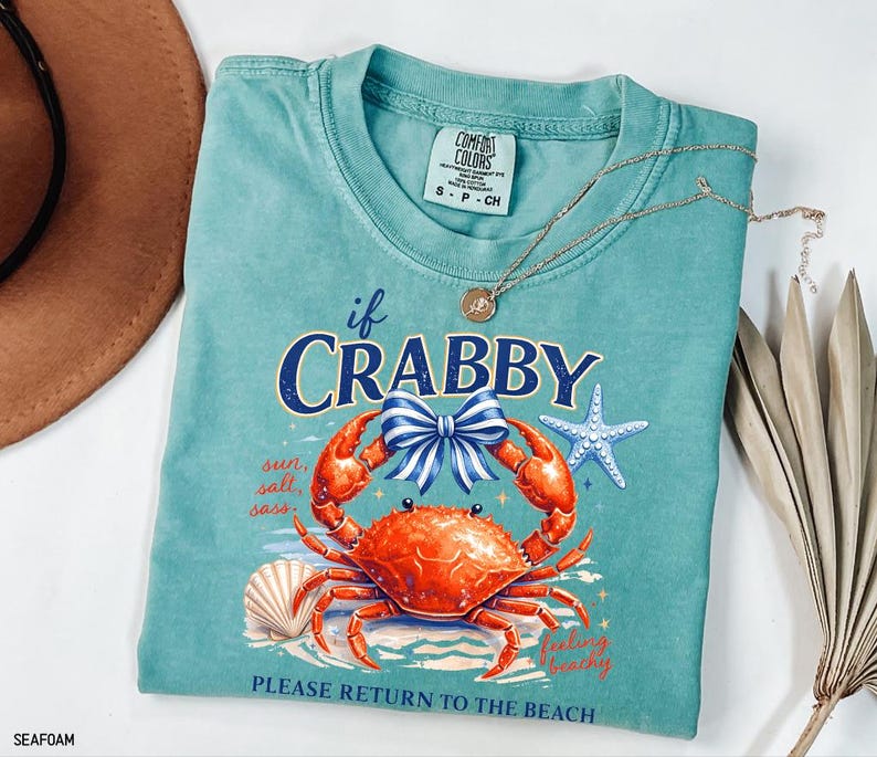 May include: Seafoam green t-shirt with a crab graphic and the text "If Crabby, Please Return to the Beach." The graphic includes a crab, bow, starfish, and seashell. A gold necklace is draped across the shirt.