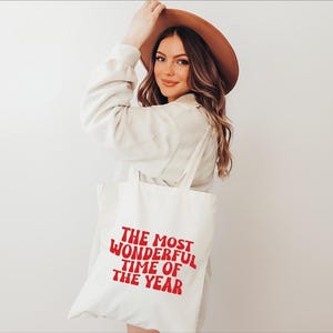 May include: A white canvas tote bag featuring the red text "THE MOST WONDERFUL TIME OF THE YEAR" printed in a bold, wavy font. The bag has two shoulder straps and is constructed from a plain, light-colored fabric material with a simple design.