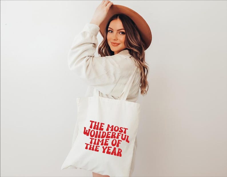 May include: A white canvas tote bag featuring the red text "THE MOST WONDERFUL TIME OF THE YEAR" printed in a bold, wavy font. The bag has two shoulder straps and is constructed from a plain, light-colored fabric material with a simple design.