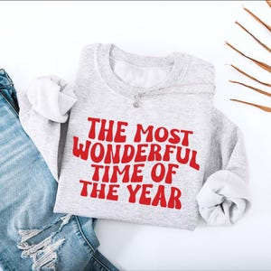 May include: A light gray crewneck sweatshirt featuring the red text THE MOST WONDERFUL TIME OF THE YEAR in a wavy font, paired with distressed blue denim jeans. The sweatshirt has long sleeves and a ribbed collar, cuffs, and hemline.