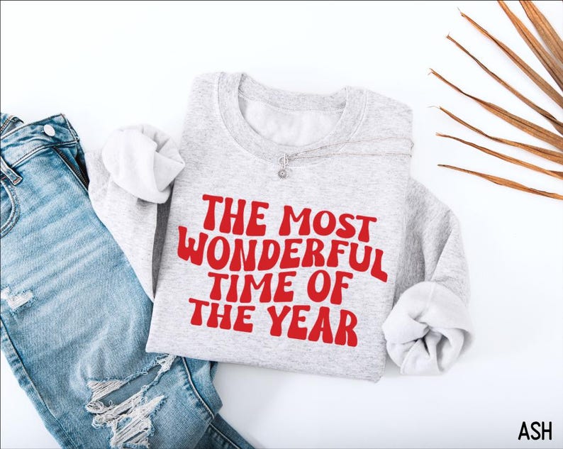 May include: A light gray crewneck sweatshirt featuring the red text THE MOST WONDERFUL TIME OF THE YEAR in a wavy font, paired with distressed blue denim jeans. The sweatshirt has long sleeves and a ribbed collar, cuffs, and hemline.
