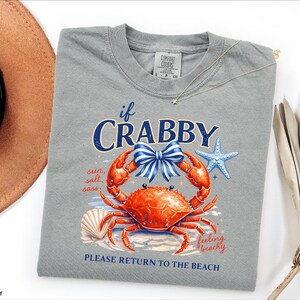 May include: A grey t-shirt with a graphic of a crab, a seashell, and a starfish. The crab is orange with a blue and white bow. Text on the shirt reads "if CRABBY" and "PLEASE RETURN TO THE BEACH". The shirt is a Comfort Colors brand.