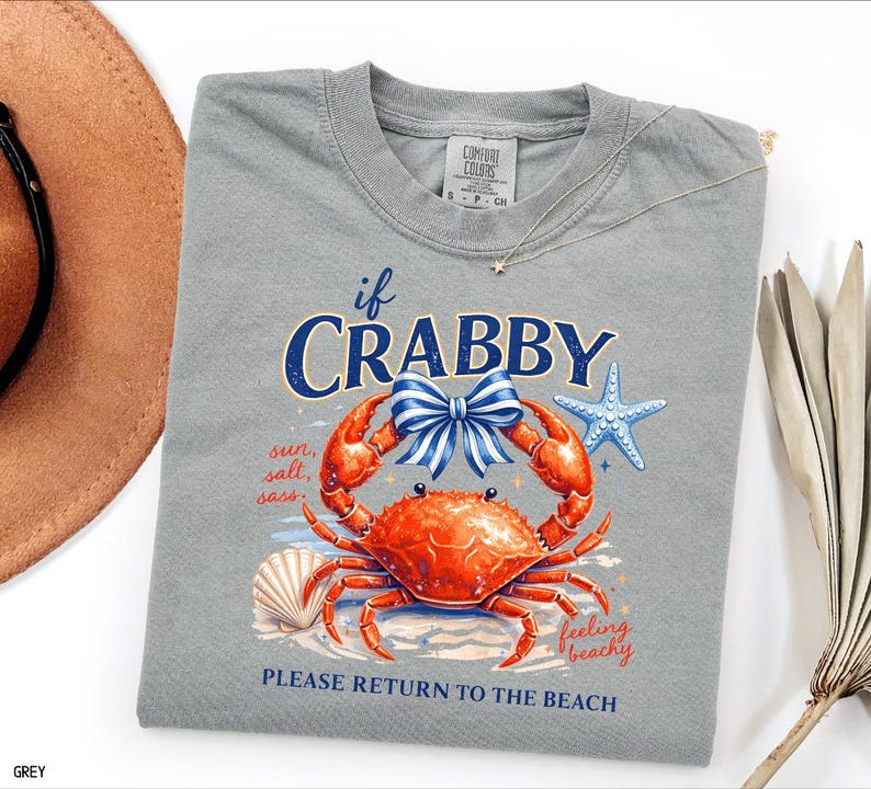 May include: A grey t-shirt with a graphic of a crab, a seashell, and a starfish. The crab is orange with a blue and white bow. Text on the shirt reads "if CRABBY" and "PLEASE RETURN TO THE BEACH". The shirt is a Comfort Colors brand.