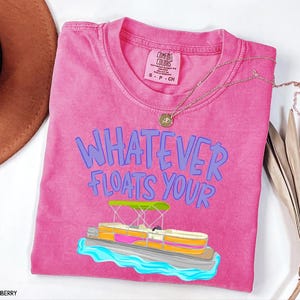 Puede incluir: Una camiseta rosa Comfort Colors con la frase azul "WHATEVER FLOATS YOUR". Un gr&aacute;fico de barco pont&oacute;n est&aacute; debajo del texto. Un collar dorado y un sombrero marr&oacute;n tambi&eacute;n est&aacute;n en la imagen.
