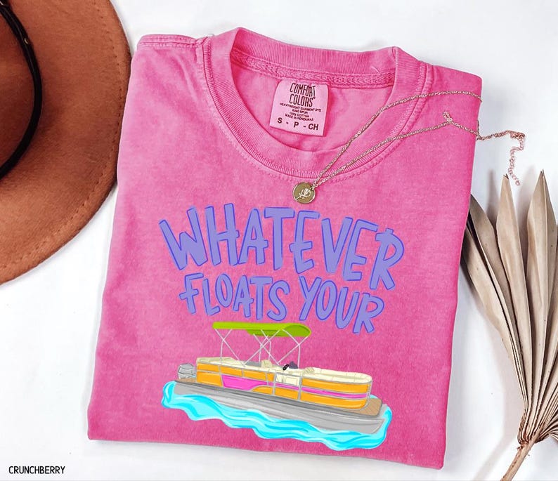 Puede incluir: Una camiseta rosa Comfort Colors con la frase azul "WHATEVER FLOATS YOUR". Un gr&aacute;fico de barco pont&oacute;n est&aacute; debajo del texto. Un collar dorado y un sombrero marr&oacute;n tambi&eacute;n est&aacute;n en la imagen.