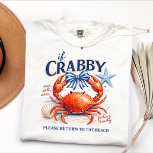 May include: White t-shirt with a crab graphic. The crab is red with a blue bow, and the text reads "if CRABBY" and "PLEASE RETURN TO THE BEACH". A starfish and seashell are also featured in the design. The shirt is a Comfort Colors brand.
