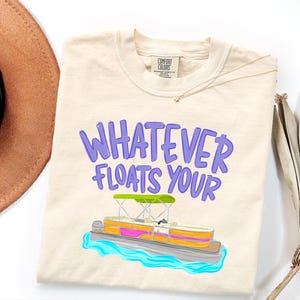 Puede incluir: Camiseta de color marfil con la frase "WHATEVER FLOATS YOUR" en letras moradas. Un dise&ntilde;o colorido de un pont&oacute;n sobre agua azul. La camiseta es de la marca Comfort Colors.