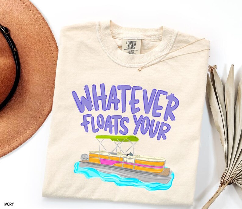 Puede incluir: Camiseta de color marfil con la frase "WHATEVER FLOATS YOUR" en letras moradas. Un dise&ntilde;o colorido de un pont&oacute;n sobre agua azul. La camiseta es de la marca Comfort Colors.