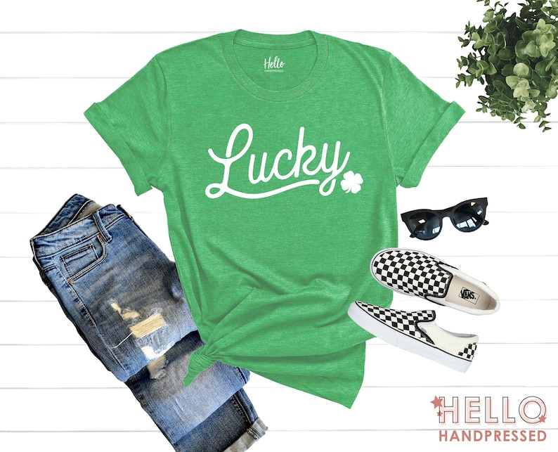 K&ouml;nnte beinhalten: Gr&uuml;nes T-Shirt mit dem Wort "Lucky" in wei&szlig;er Kursivschrift und einem Kleeblatt. Das Shirt wird mit einer blauen Jeans, karierten Slipper und einer Sonnenbrille kombiniert.