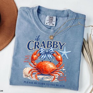 May include: A blue jean Comfort Colors t-shirt with a crab graphic. The graphic includes a crab with a blue bow, a starfish, and the text "if Crabby, Please Return to the Beach". A gold necklace is draped across the shirt.