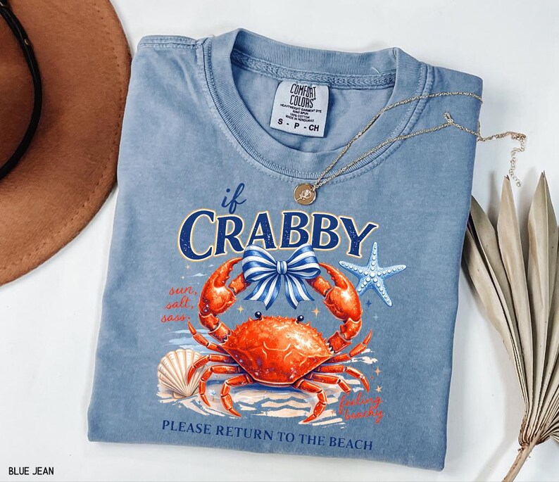 May include: A blue jean Comfort Colors t-shirt with a crab graphic. The graphic includes a crab with a blue bow, a starfish, and the text "if Crabby, Please Return to the Beach". A gold necklace is draped across the shirt.
