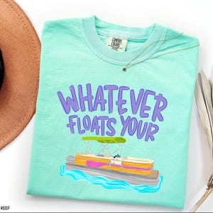 Puede incluir: Una camiseta Comfort Colors de color turquesa claro con la frase "WHATEVER FLOATS YOUR" en letras moradas. Debajo del texto hay una colorida ilustraci&oacute;n de un barco pont&oacute;n en el agua. Se ve un sombrero marr&oacute;n y un collar dorado.
