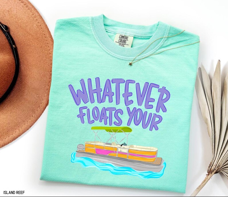 Puede incluir: Una camiseta Comfort Colors de color turquesa claro con la frase "WHATEVER FLOATS YOUR" en letras moradas. Debajo del texto hay una colorida ilustraci&oacute;n de un barco pont&oacute;n en el agua. Se ve un sombrero marr&oacute;n y un collar dorado.