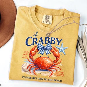 May include: Mustard-colored t-shirt with a crab graphic and the text "If Crabby, Please Return to the Beach." The design includes a red crab, blue bow, starfish, and seashell. A silver necklace is draped across the shirt.