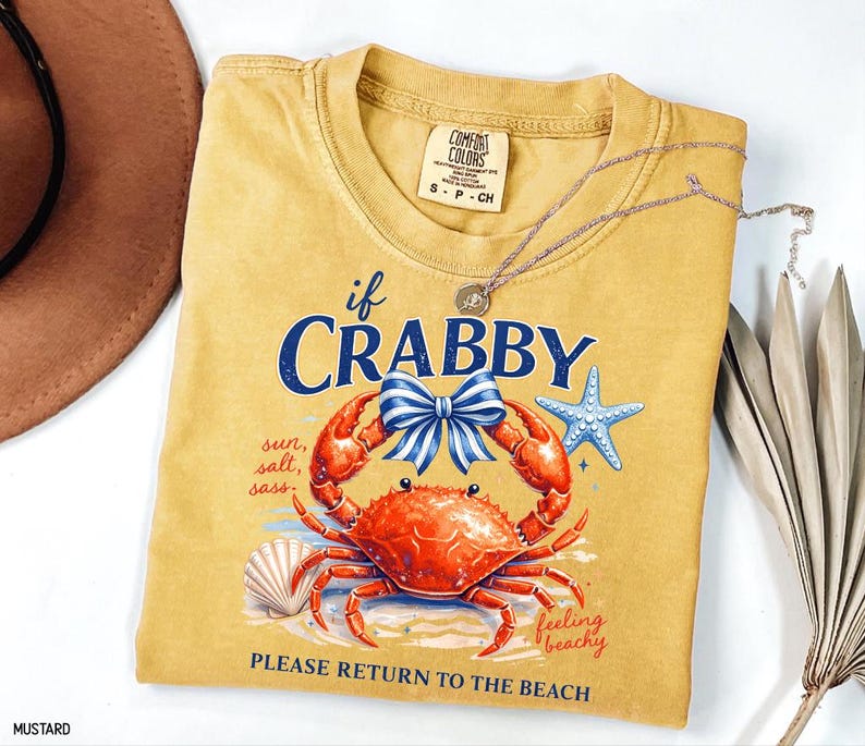 May include: Mustard-colored t-shirt with a crab graphic and the text "If Crabby, Please Return to the Beach." The design includes a red crab, blue bow, starfish, and seashell. A silver necklace is draped across the shirt.