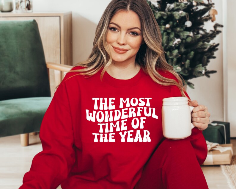 May include: A bright red crewneck sweatshirt featuring the white text "THE MOST WONDERFUL TIME OF THE YEAR" in a bold, wavy font. The garment has long sleeves and a ribbed neckline, providing a classic fit for the holiday season.