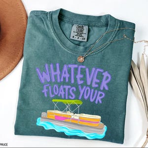 Puede incluir: Camiseta azul abeto con la frase "WHATEVER FLOATS YOUR" en letras moradas. Un gr&aacute;fico de un pont&oacute;n est&aacute; debajo del texto. La camiseta tiene cuello redondo y mangas cortas.