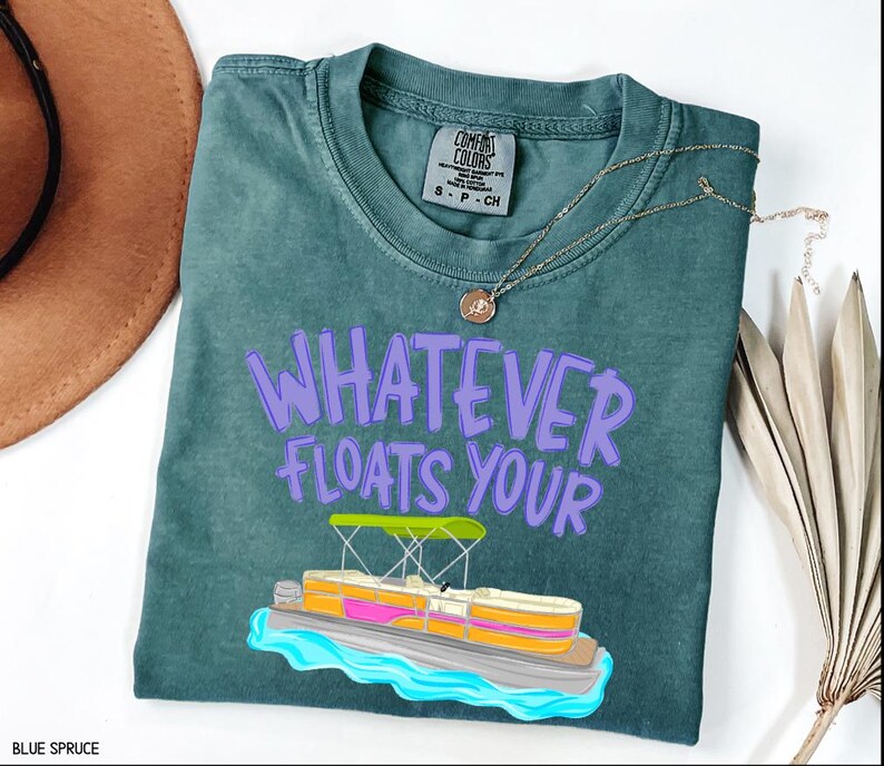Puede incluir: Camiseta azul abeto con la frase "WHATEVER FLOATS YOUR" en letras moradas. Un gr&aacute;fico de un pont&oacute;n est&aacute; debajo del texto. La camiseta tiene cuello redondo y mangas cortas.