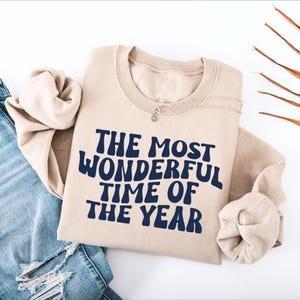 May include: A sand-colored crewneck sweatshirt featuring the dark navy blue text THE MOST WONDERFUL TIME OF THE YEAR printed in a bold, wavy font across the chest. The garment is paired with light blue distressed denim jeans.