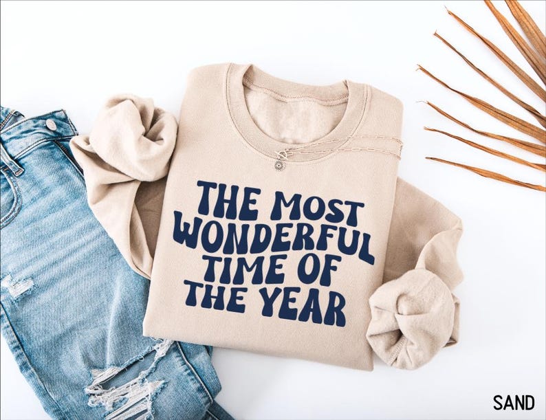 May include: A sand-colored crewneck sweatshirt featuring the dark navy blue text THE MOST WONDERFUL TIME OF THE YEAR printed in a bold, wavy font across the chest. The garment is paired with light blue distressed denim jeans.